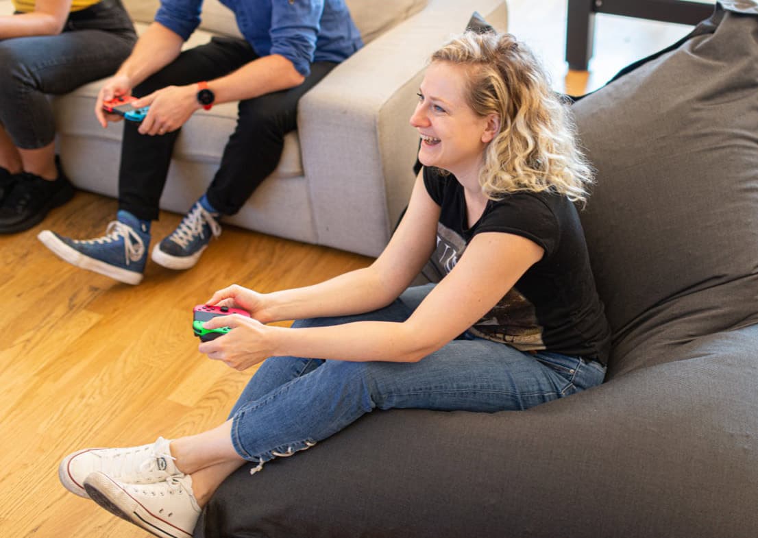 People playing video games with colourful controllers while sitting on grey sofas in a living room with wooden flooring.