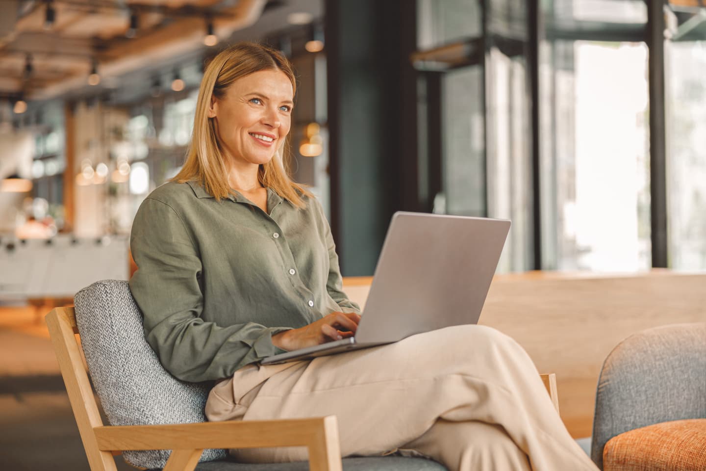 Woman in green shirt smiling while working on laptop in modern office space with large windows.