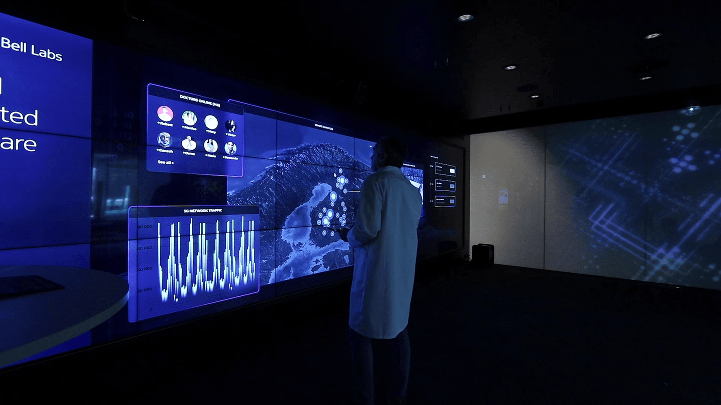 Person in lab coat viewing Bell Labs digital displays showing 5G network traffic and geographical data in blue lighting.