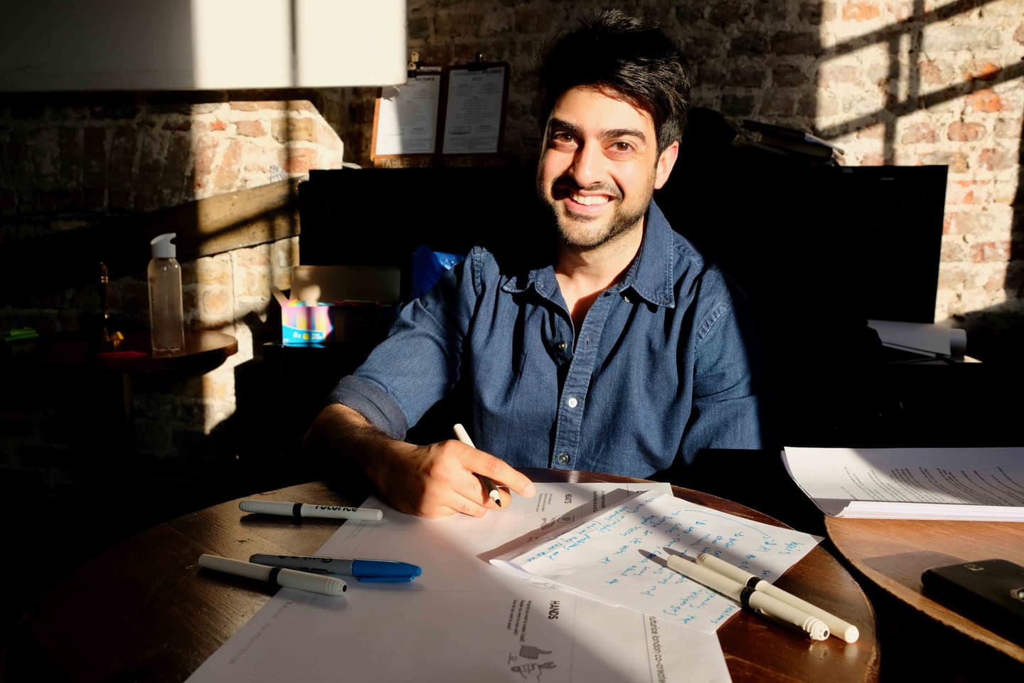 Person in blue denim shirt smiling while writing notes at wooden table in rustic brick-walled café or workspace.