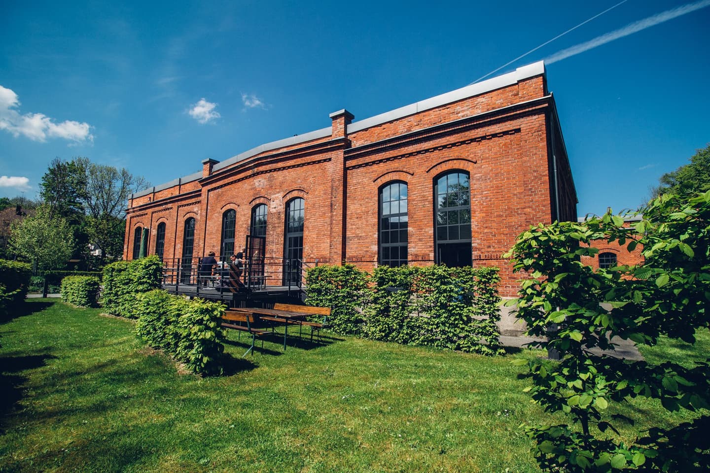 Historic red brick building with arched windows surrounded by green lawn and shrubs under a bright blue sky.